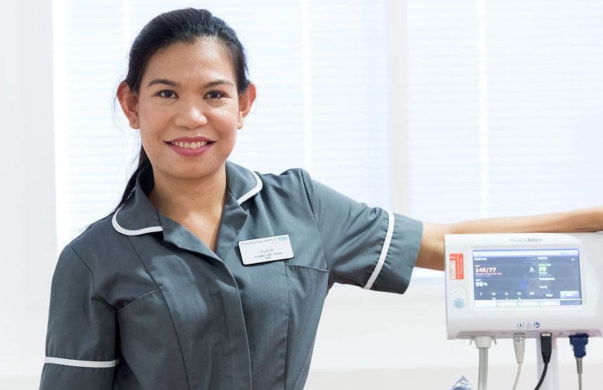 A smiling healthcare assistant in a grey uniform standing beside a vital-signs monitor in a bright clinical room