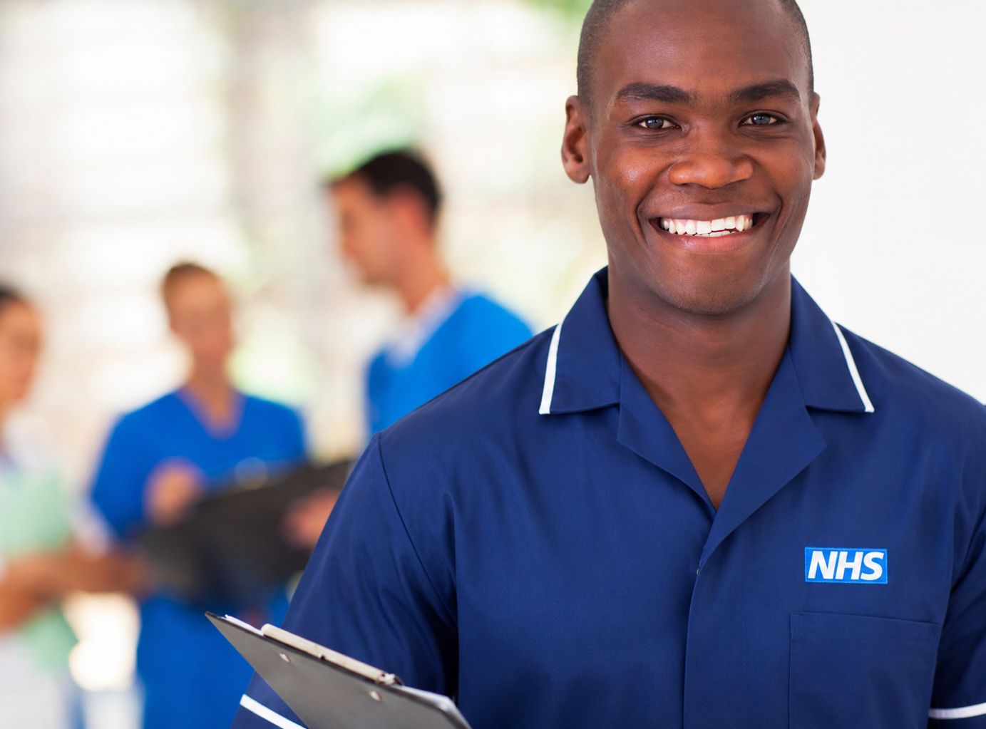 A smiling NHS healthcare worker in a navy tunic holding a clipboard, with colleagues in the background
