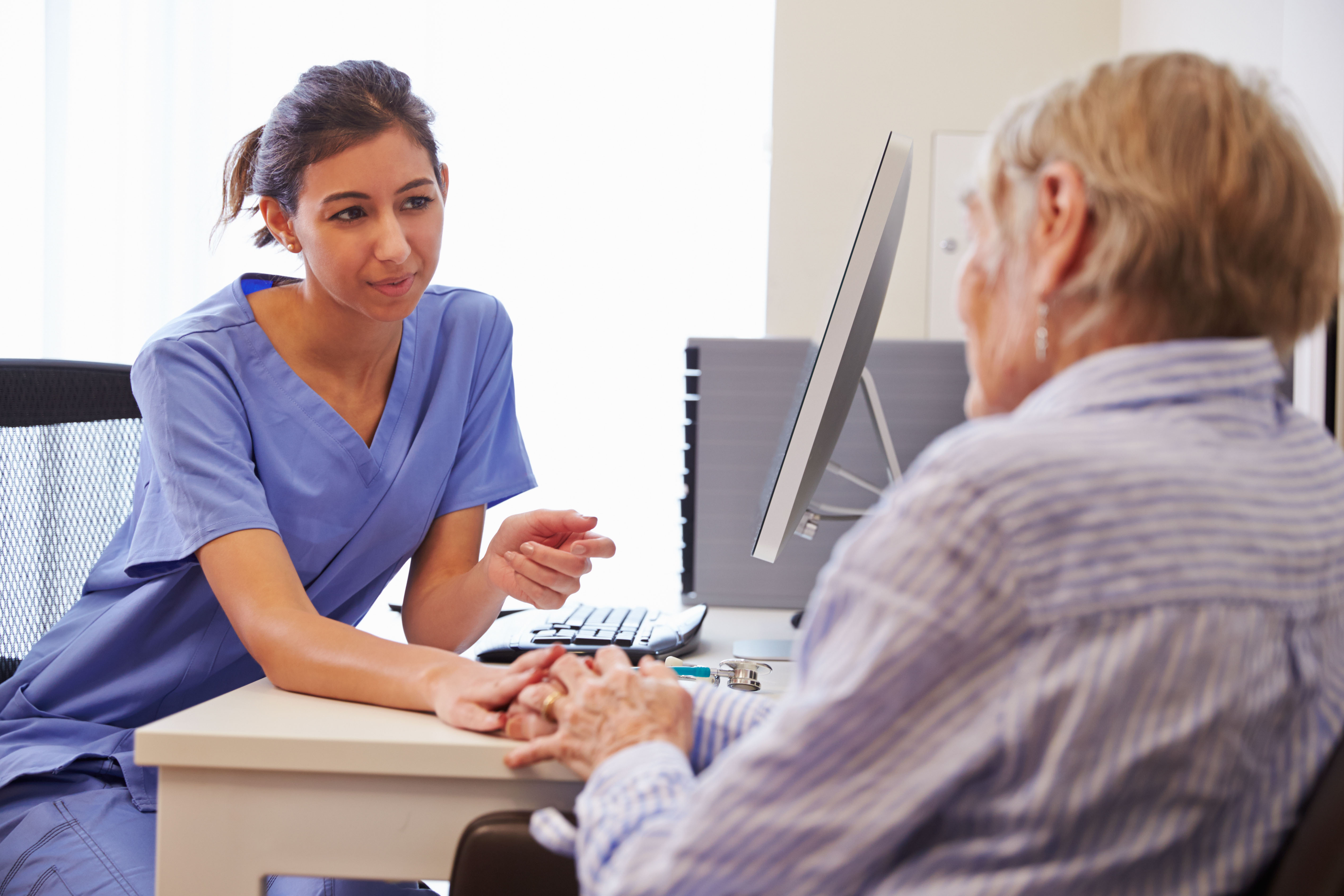 A nurse in blue scrubs sitting at a desk, holding hands and talking compassionately with an elderly patient